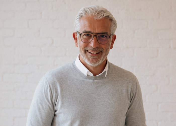 Smiling older man with gray hair and glasses wearing a light gray sweater and brown pants stands against a white brick wall.