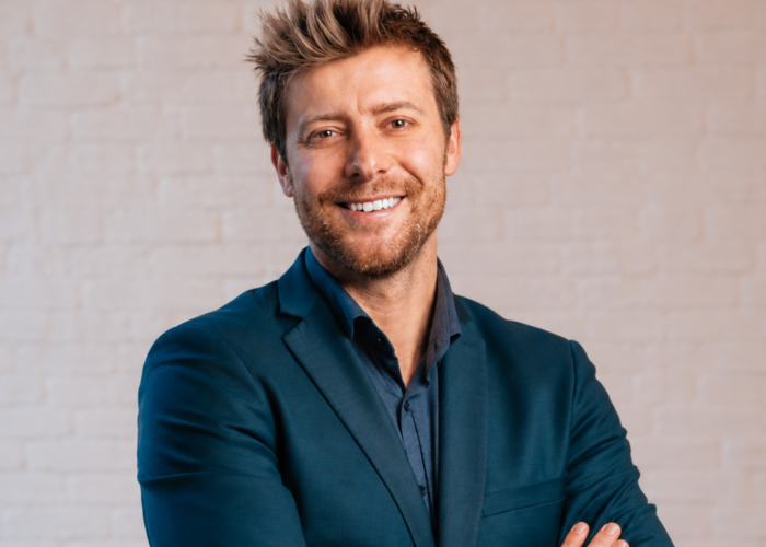 Smiling man in a dark blazer and shirt, standing with arms crossed against a light brick background.