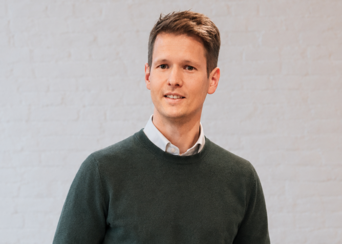 A young man in a green sweater and light shirt stands confidently against a neutral brick wall.