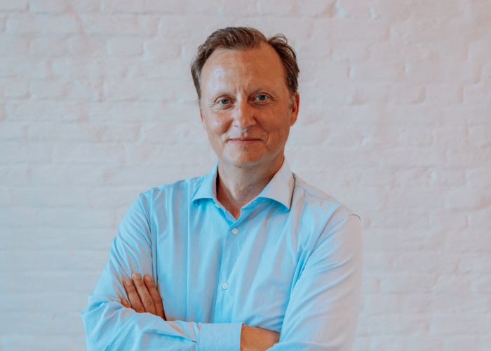 A man in a light blue shirt stands confidently with arms crossed against a white brick wall.