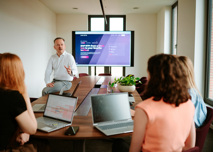 A man presents in a meeting room, while others listen with laptops open, displaying a website on a large screen.
