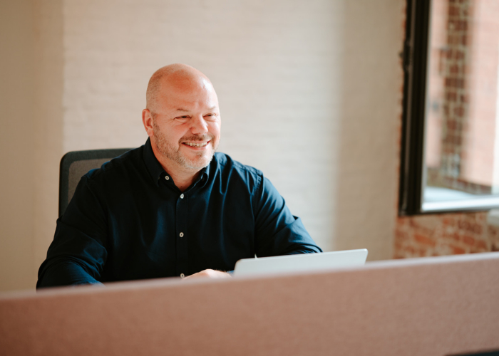 A smiling man in a dark shirt sits at a desk with a laptop, in a bright office space with large windows.