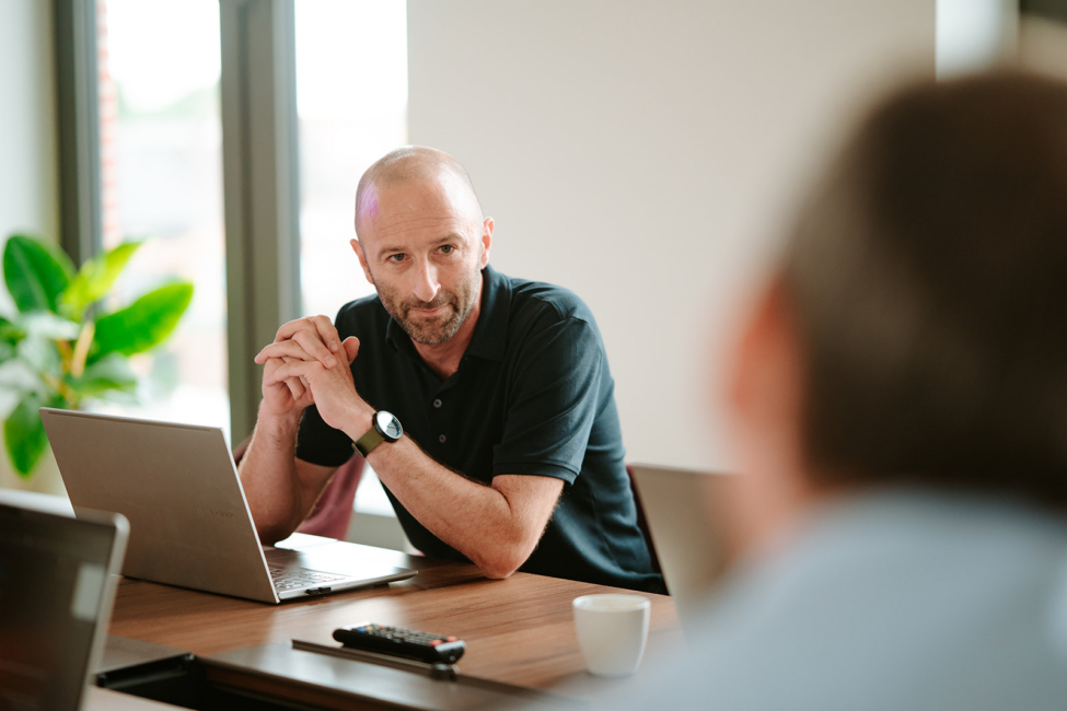A man with a bald head and beard sits at a conference table, looking engaged in conversation, with laptops and a plant visible.