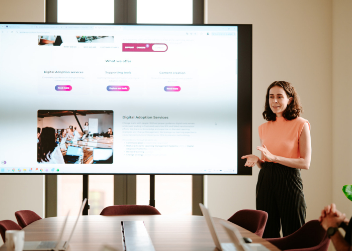 A woman in an orange top presents digital adoption services to an audience in a modern meeting room.