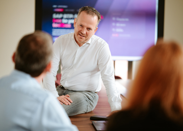 A man in a white shirt speaks to two people in a meeting room with a screen displaying information behind him.