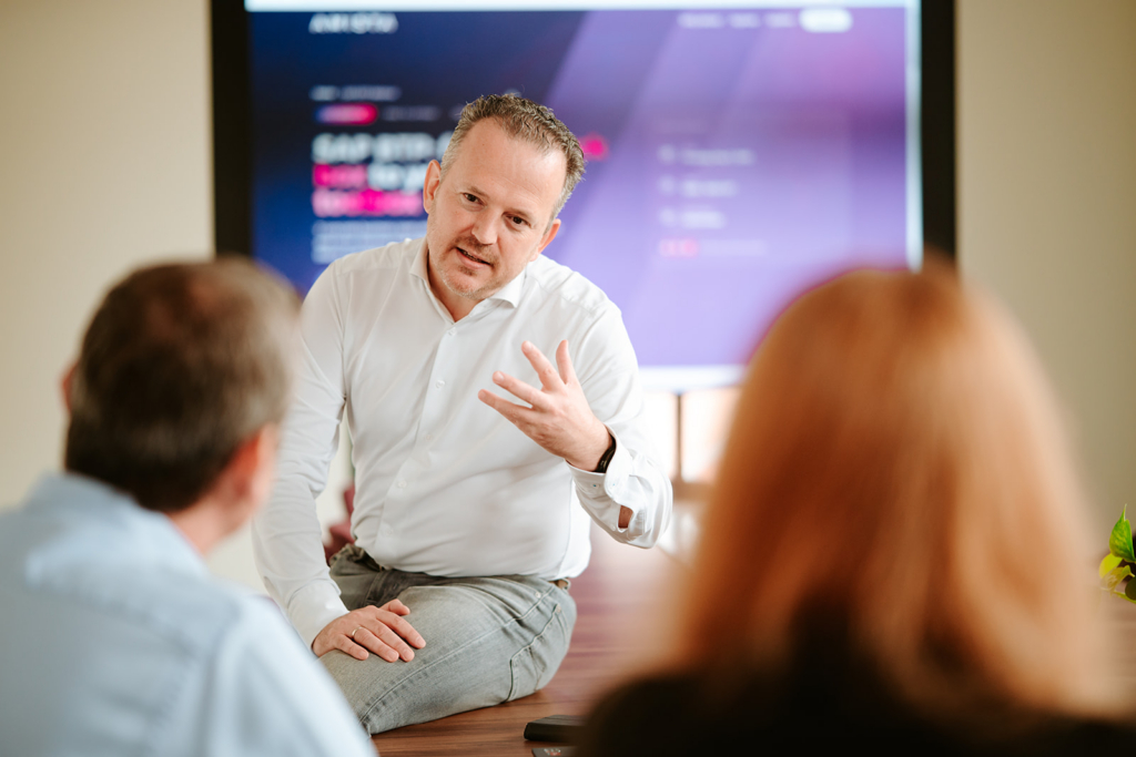 A person in a white shirt discusses with two individuals in a meeting room, a screen displays information in the background.