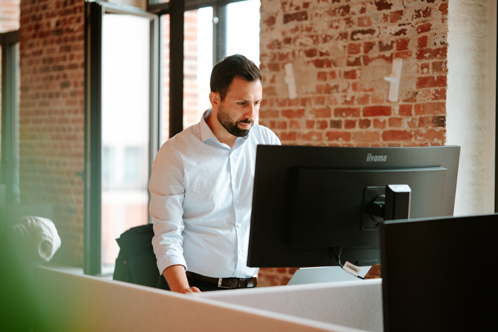 Man in a light blue shirt stands at a desk, focused on a computer monitor in a bright, modern office space.