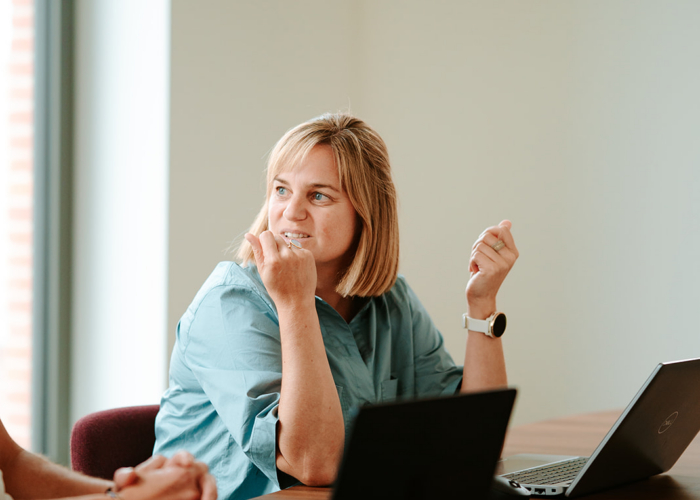 A woman in a blue shirt sits at a table, engaging in conversation with laptops open in front of her.