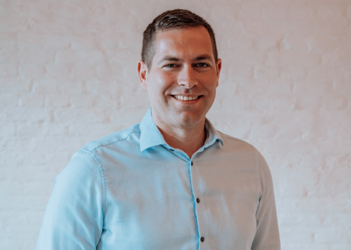 Smiling man in a light blue shirt standing in front of a plain white wall.