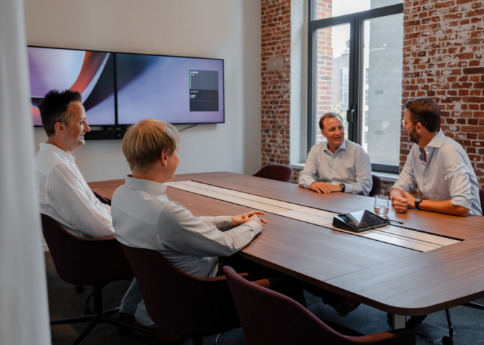 Four professionals engage in discussion around a conference table, with monitors displaying content in a modern meeting room.