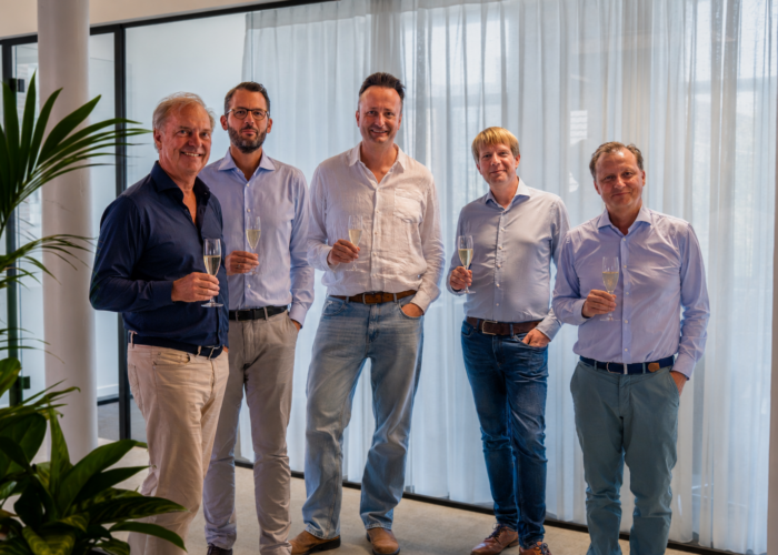 Five men stand together, holding champagne glasses, in a bright, modern interior with sheer curtains.