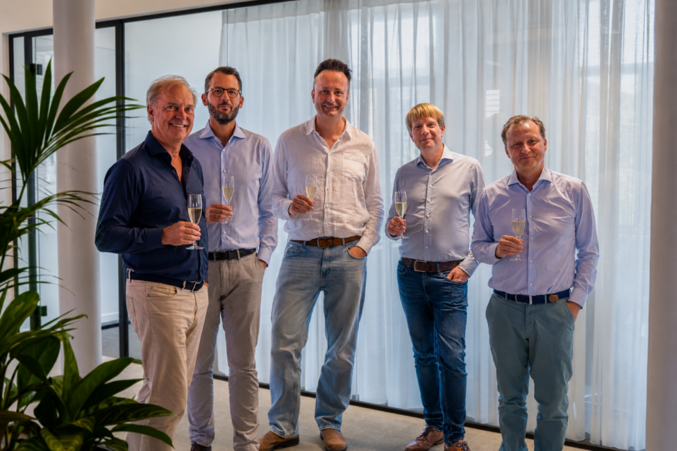 Five men stand together, holding champagne glasses, in a bright, modern interior with sheer curtains.