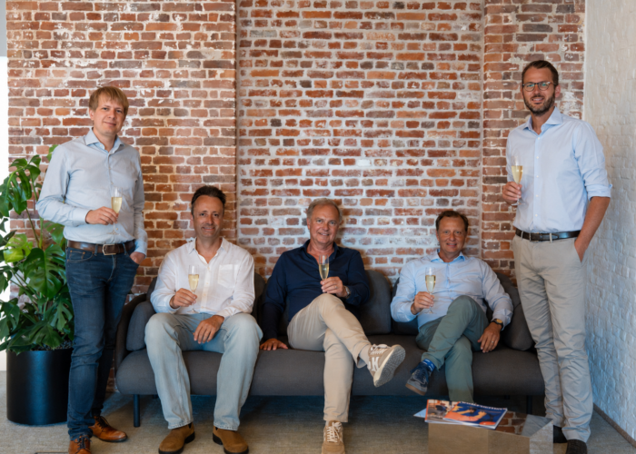 Five men holding champagne glasses pose on a sofa against a brick wall, smiling in a bright, modern setting.
