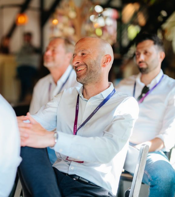 A smiling man in a light shirt sits among others, engaged and enjoying the moment in a bright, casual setting.