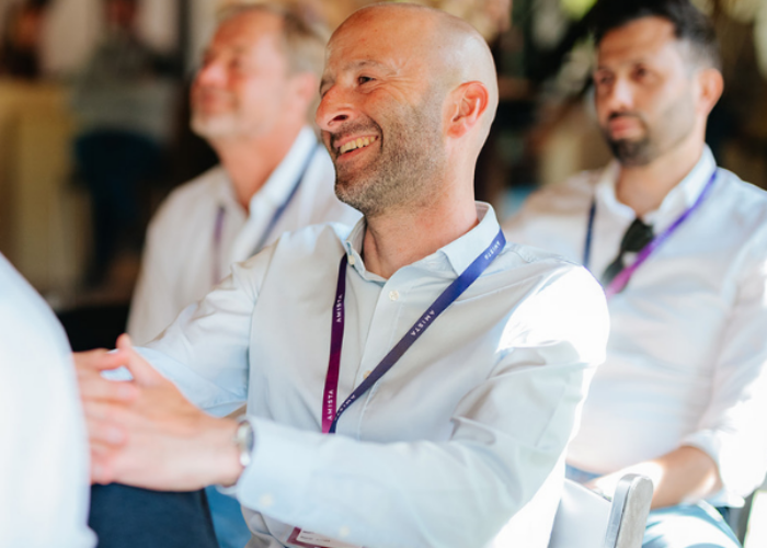 A smiling man in a light shirt sits among others, engaged and enjoying the moment in a bright, casual setting.
