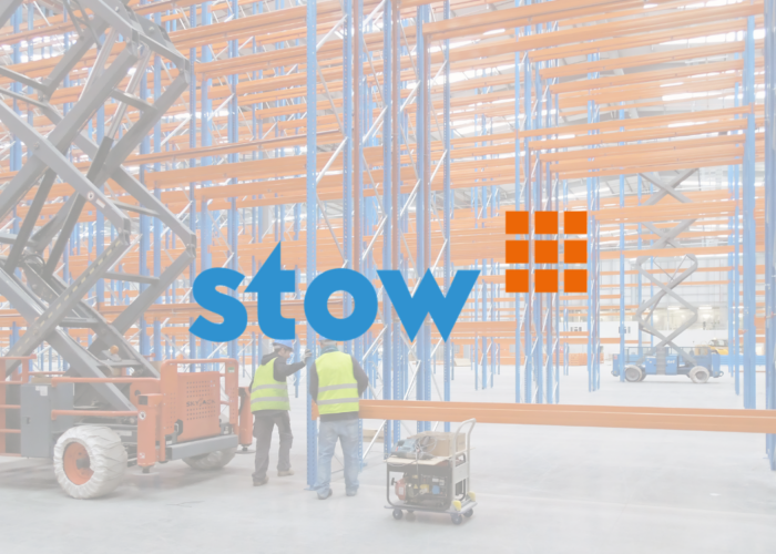 Warehouse interior with empty blue and orange storage racks, two workers in vests, and scissor lifts.